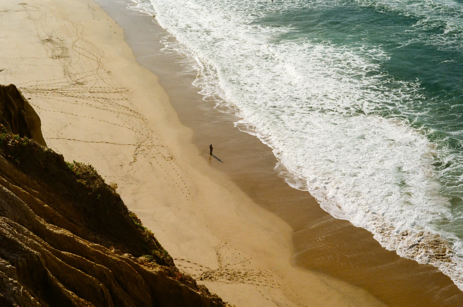 A lone person on a sandy beach beside foaming waves, viewed from a cliff.