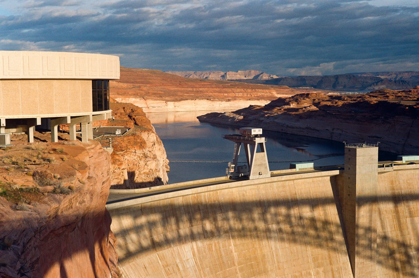 A dam and reservoir between sunlit sandstone cliffs under dark clouds.
