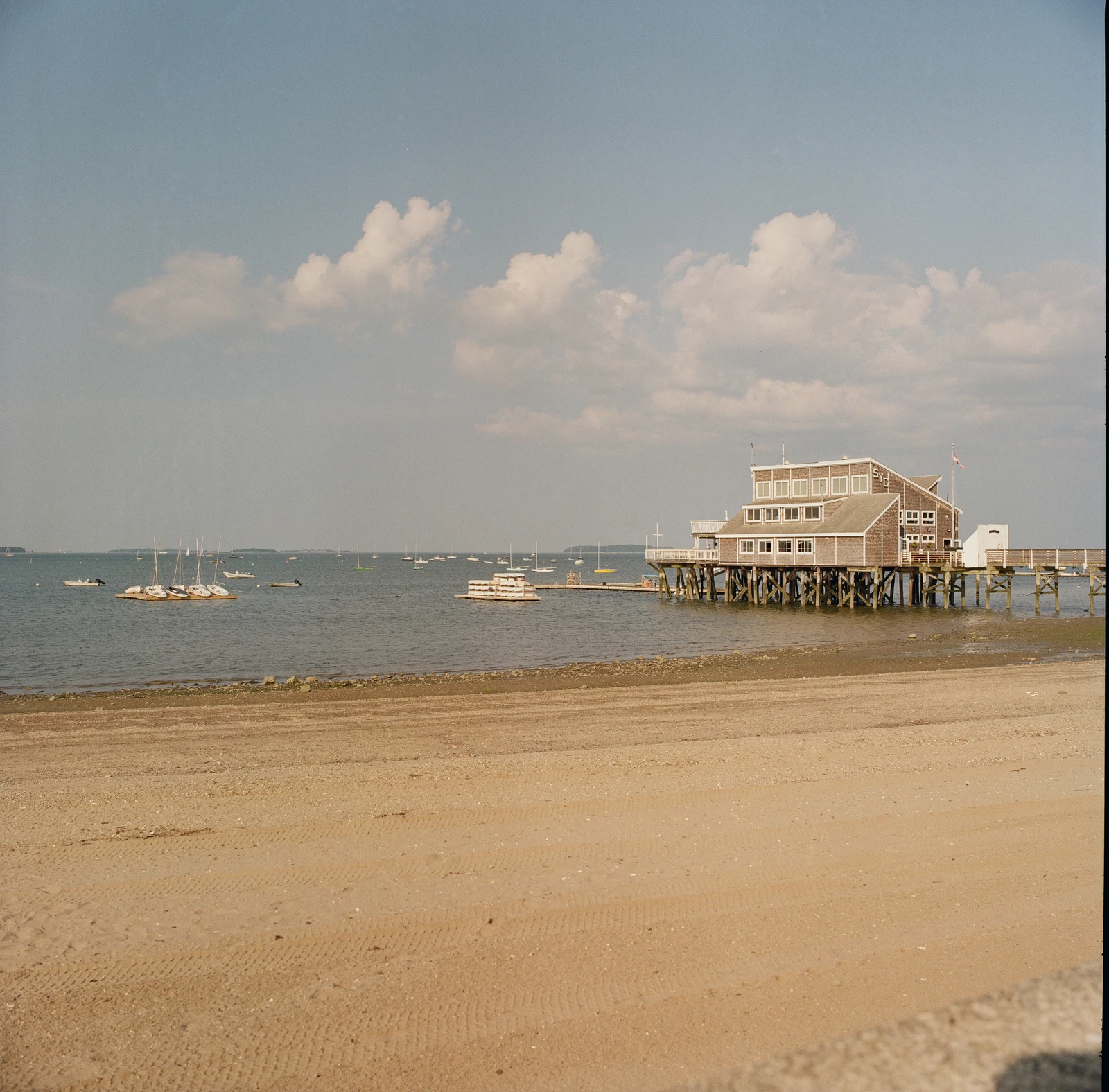 A wooden building on a coastal pier with sailboats and a sandy beach.