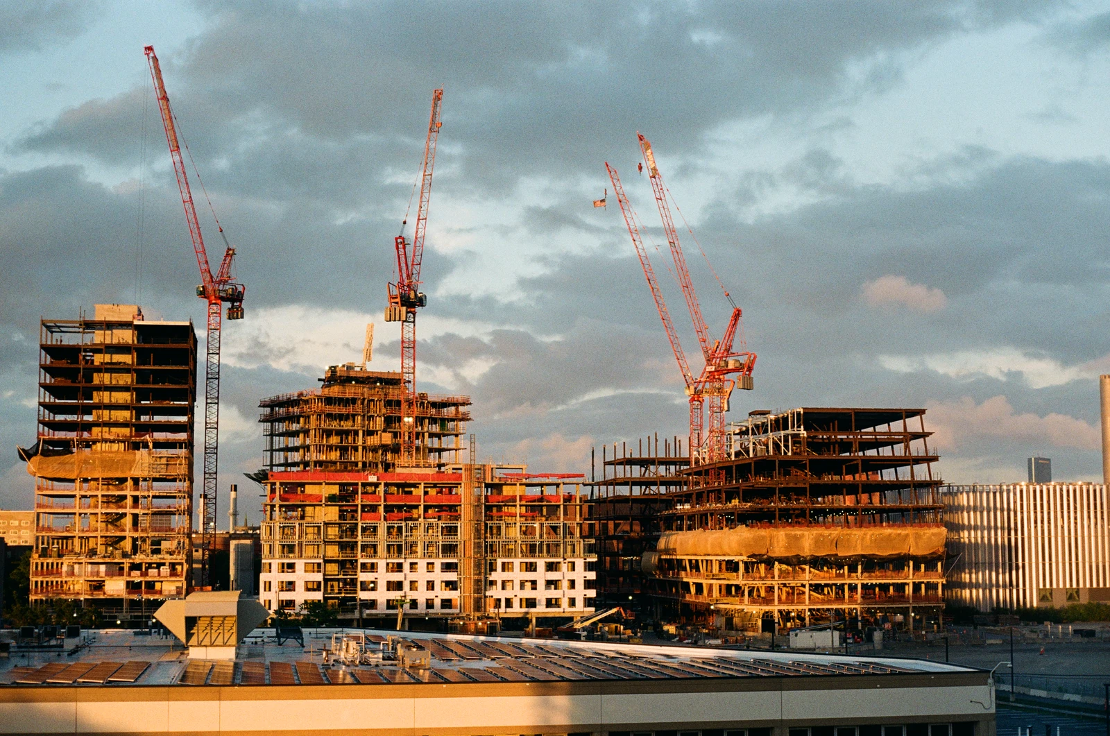 Red construction cranes and unfinished structures in warm light beneath a cloudy sky.
