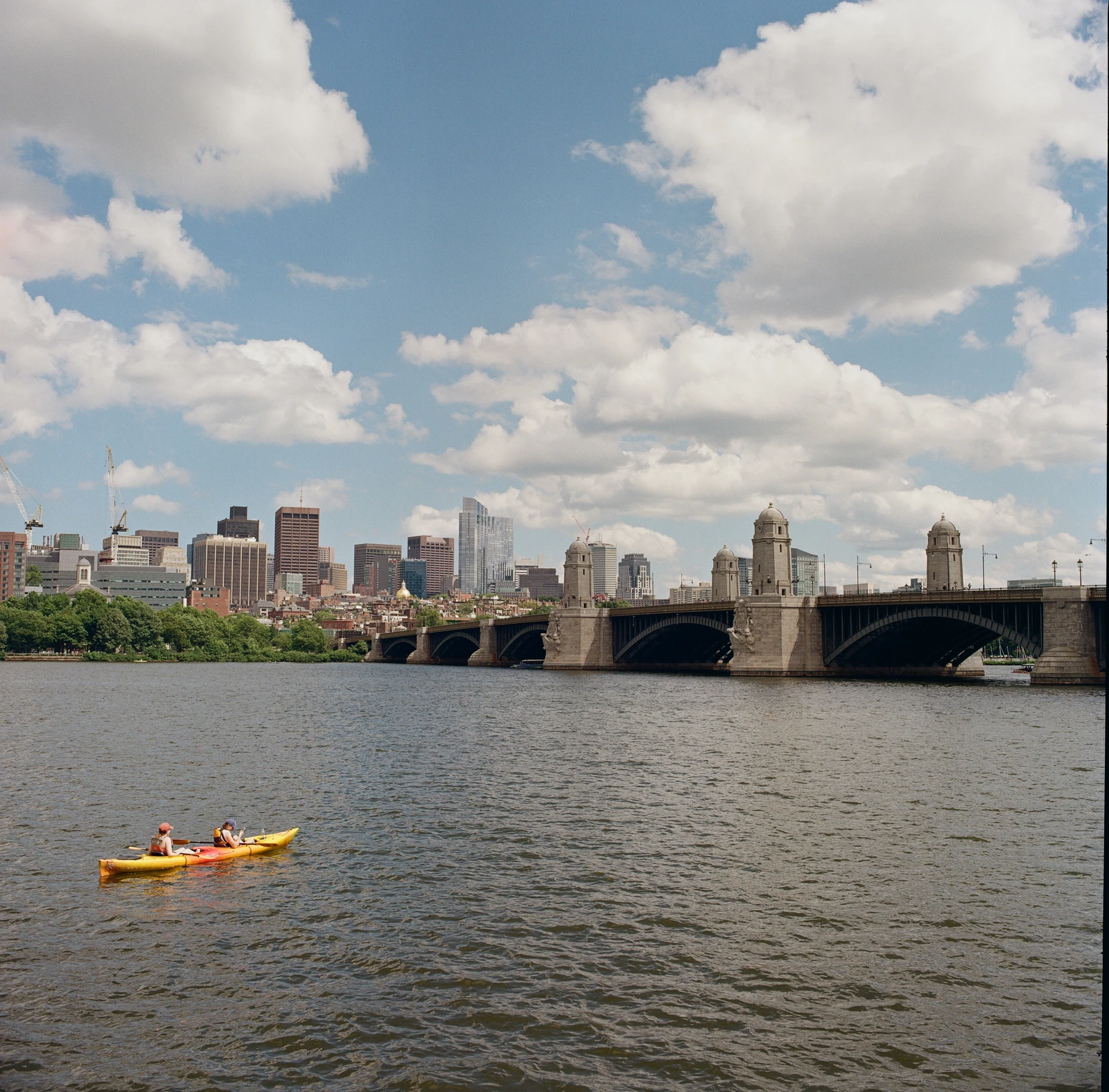 Kayakers on a river beside an arched bridge and city skyline beneath large white clouds.